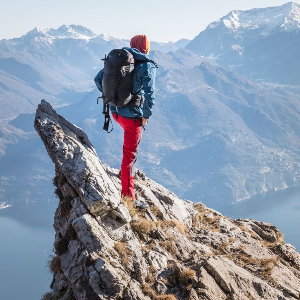 Person with a backpack standing on a mountain peak with a scenic view of mountains in the background. Mountain Clothing Collection inspiration by Natura Nation.