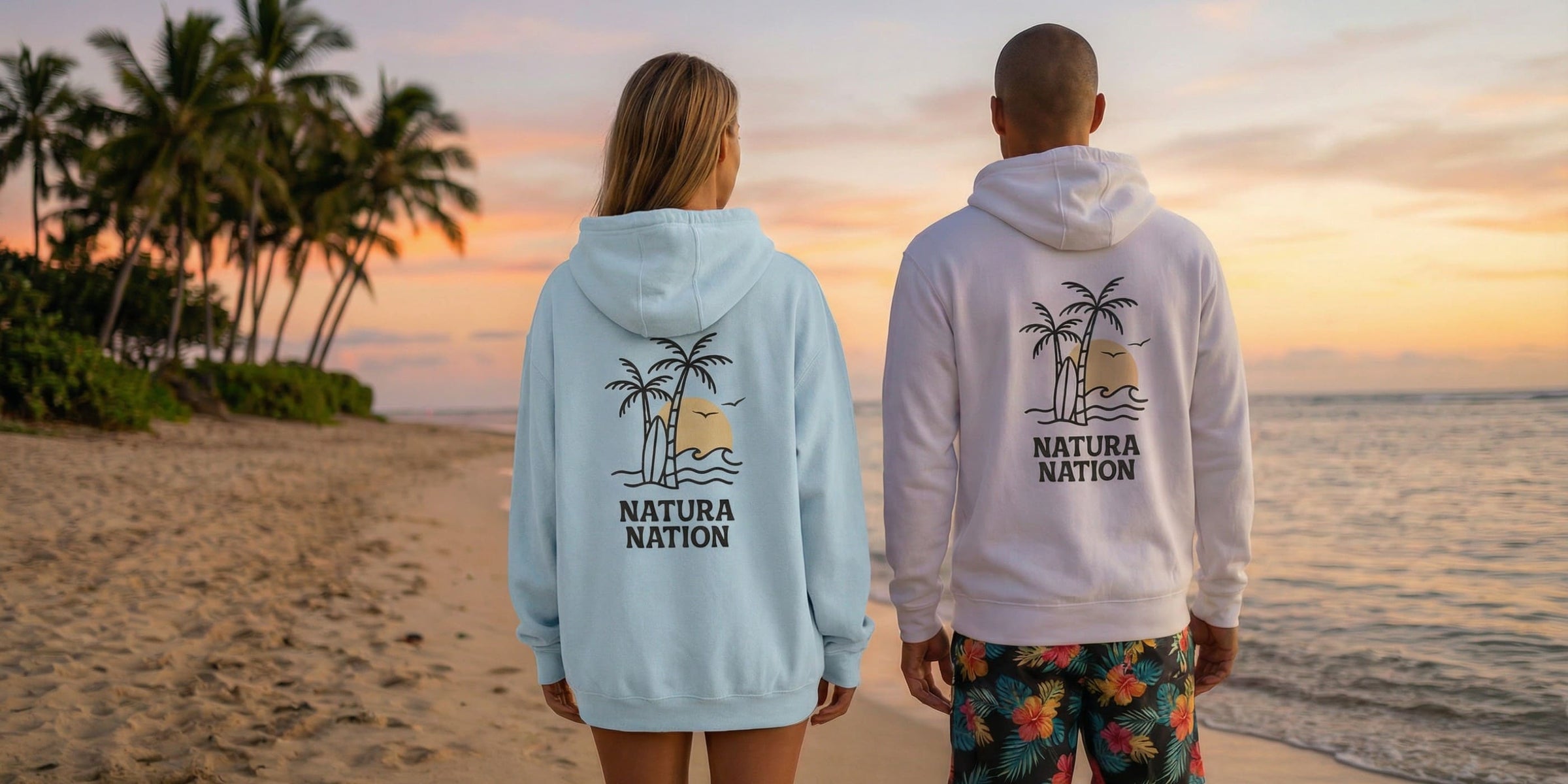 Two people wearing tropical hoodies with the 'Natura Nation' brand design on the back; they're on a beach at sunset.