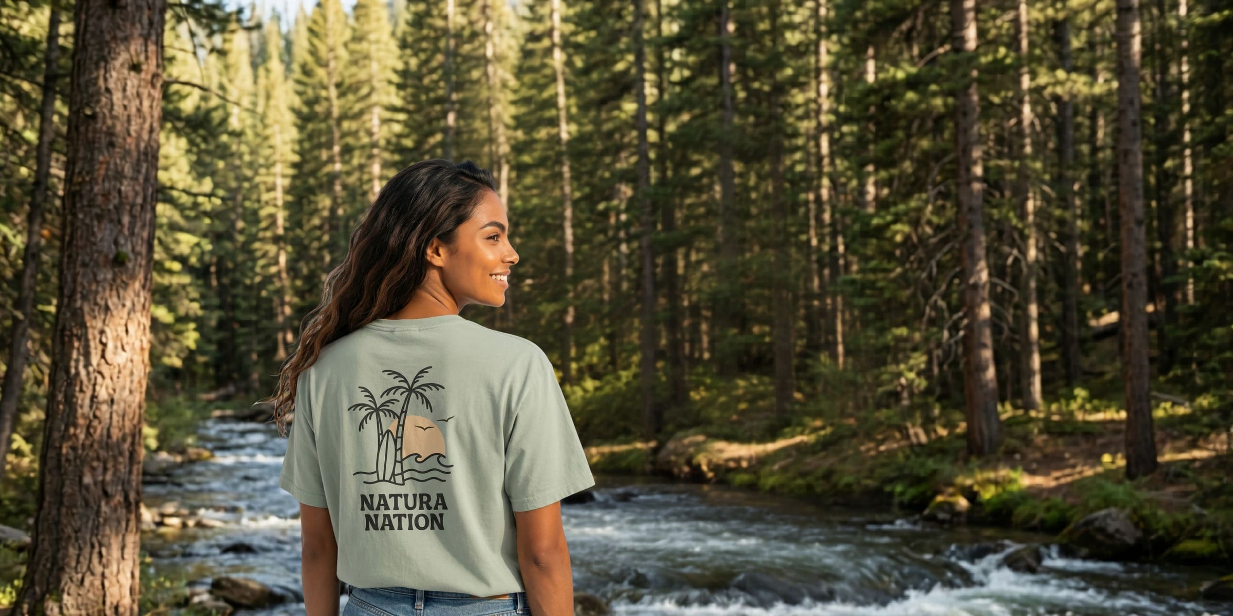 Women's T-Shirts -  A woman wearing a bay-colored nature-inspired t-shirt with 'Natura Nation' branding on the back, standing in a dense pine forest with a river in the background.