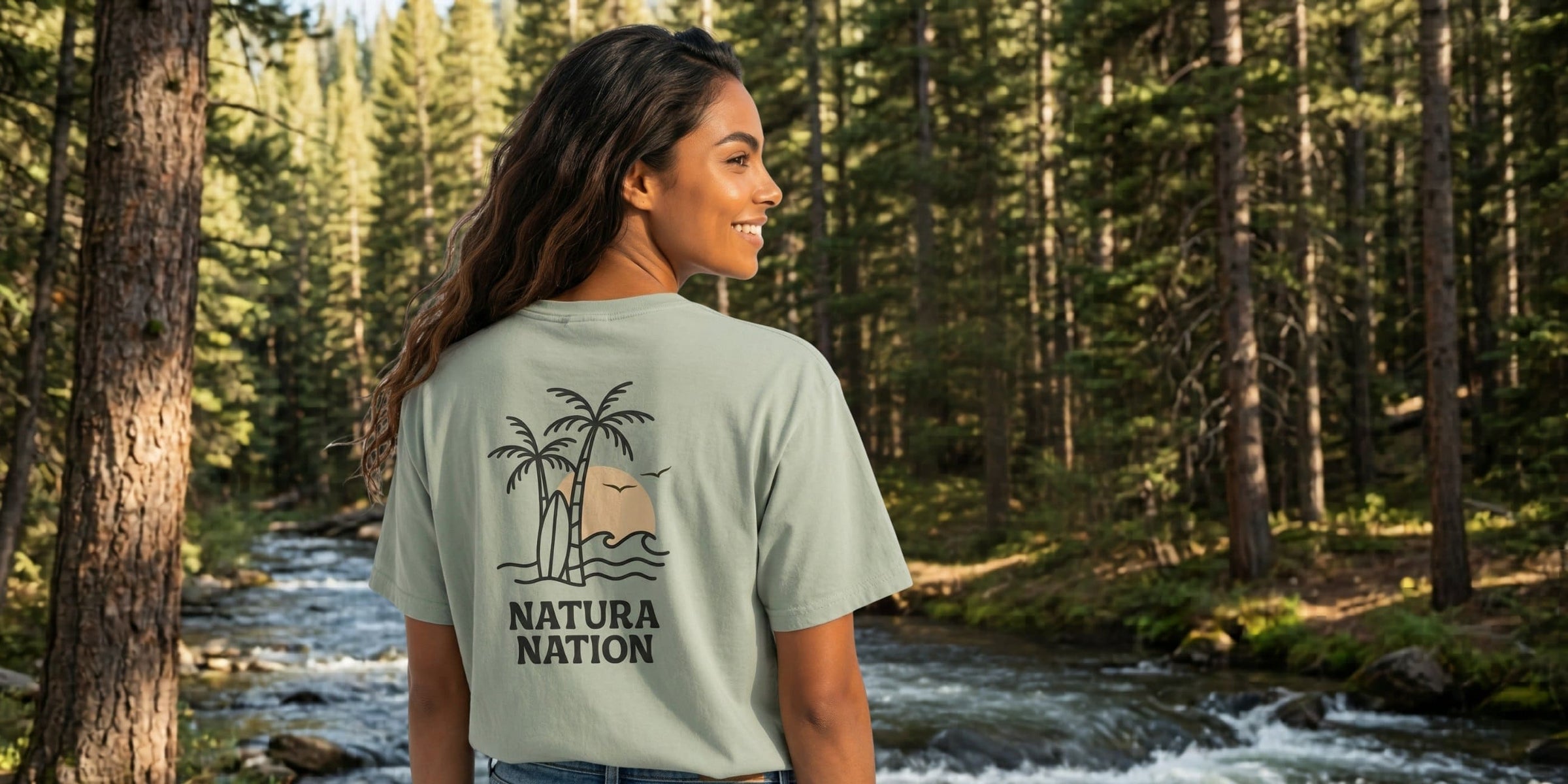 Women's T-Shirts - A woman wearing a bay-colored, nature-inspired t-shirt with 'Natura Nation' branding printed on the back, standing in a dense pine forest with a river in the background. 